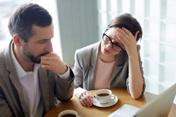 Two people sitting together at a table drinking coffee and appearing stressed while looking at a laptop screen