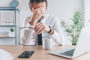 Stressed business man sitting at desk with coffee and laptop while removing glasses and rubbing brow.