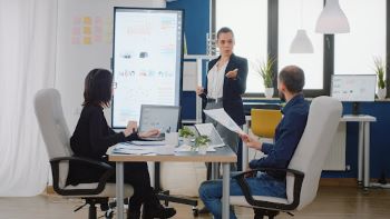 business woman standing at end of work table leading a meeting with two other business people who are using laptop computers