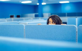 A person cautiously peering over the top of an office partition in an all-blue office environment