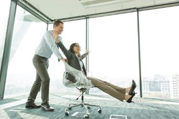 A happy woman with outstretched arms is sitting in a rolling office chair and a man is pushing her across the carpet in an empty office space with large windows looking out to a city skyline