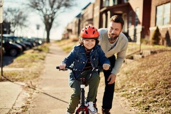 A man assisting a young girl as she rides a bike down a city sidewalk